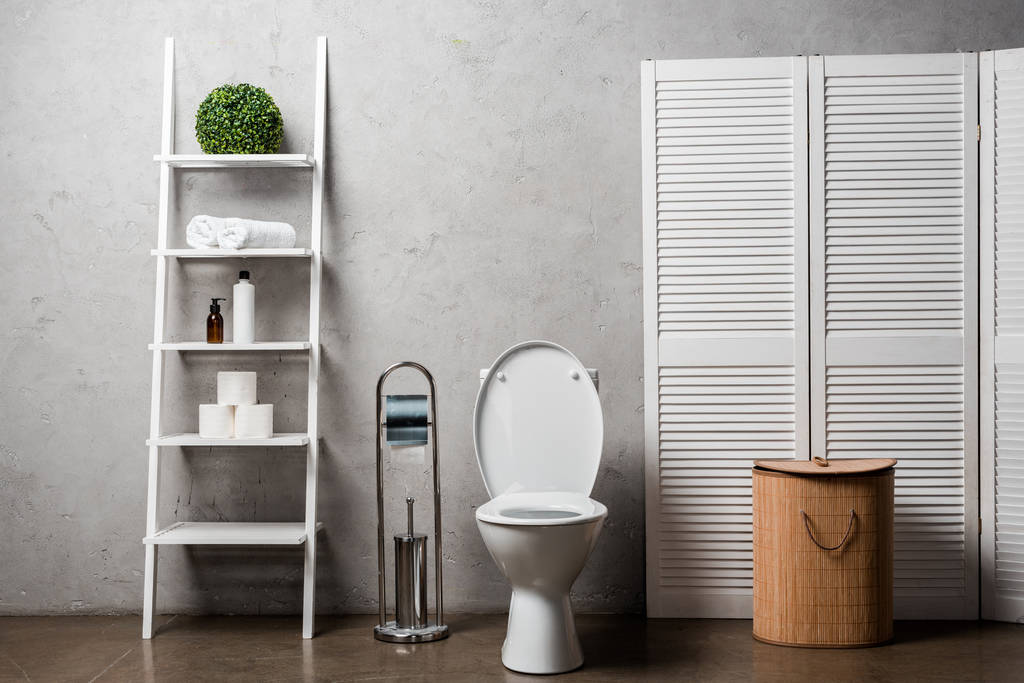 салон современной ванной комнаты с унитазом возле стойки с косметикой, полотенцами, туалетной бумагой, корзиной для белья и туалетной щеткой
 - Фото, изображение