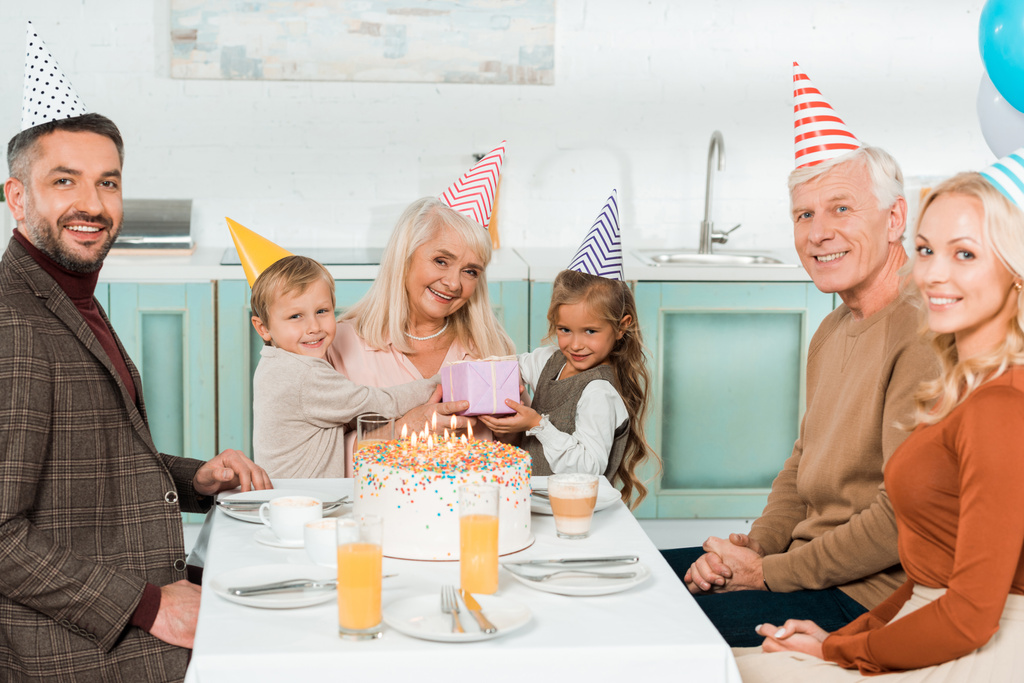 χαρούμενα παιδιά κάθονται σε γύρους της γιαγιάς, ενώ κάθεται κοντά τούρτα γενεθλίων με την οικογένεια - Φωτογραφία, εικόνα