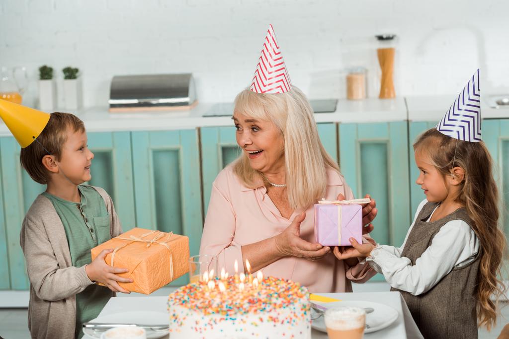 adorables niños presentando cajas de regalo a la abuela feliz sentado cerca de pastel de cumpleaños
 - Foto, imagen