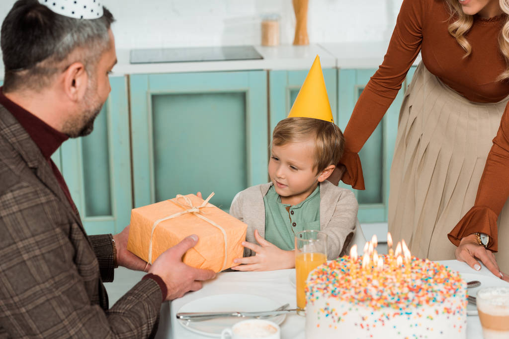 father presenting gift box to happy son while sitting at kitchen table near birthday cake - Photo, Image