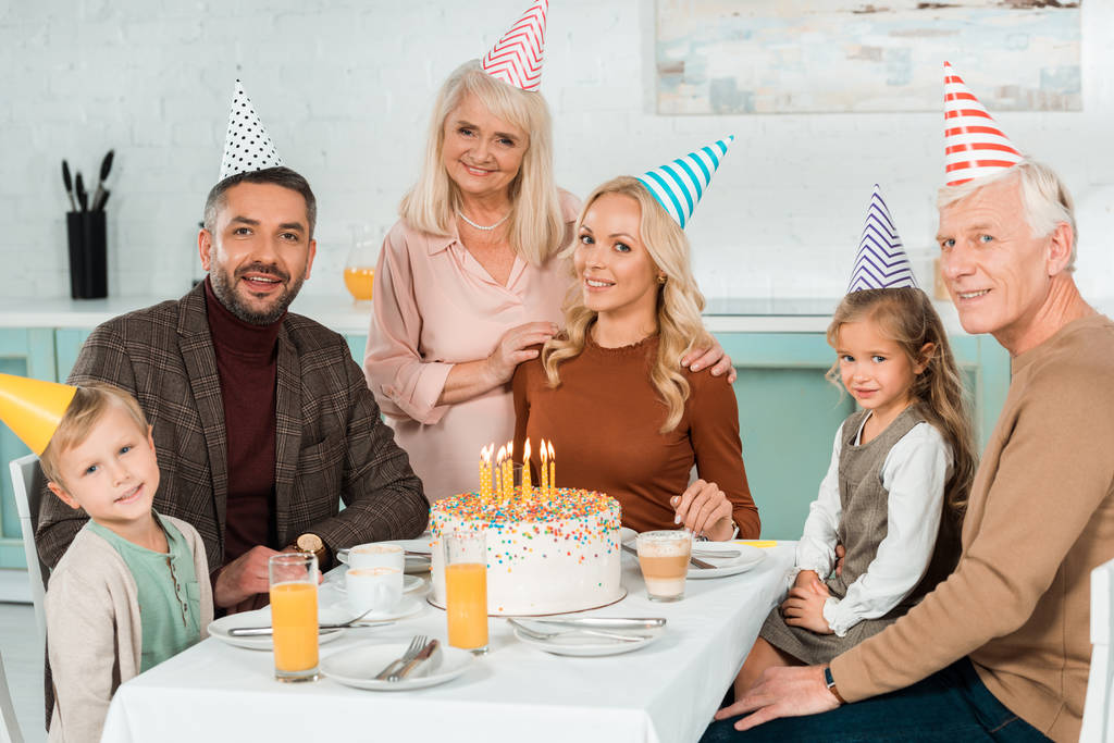 onnellinen perhe hymyilee kameran edessä istuessaan keittiön pöydässä lähellä syntymäpäiväkakkua
 - Valokuva, kuva