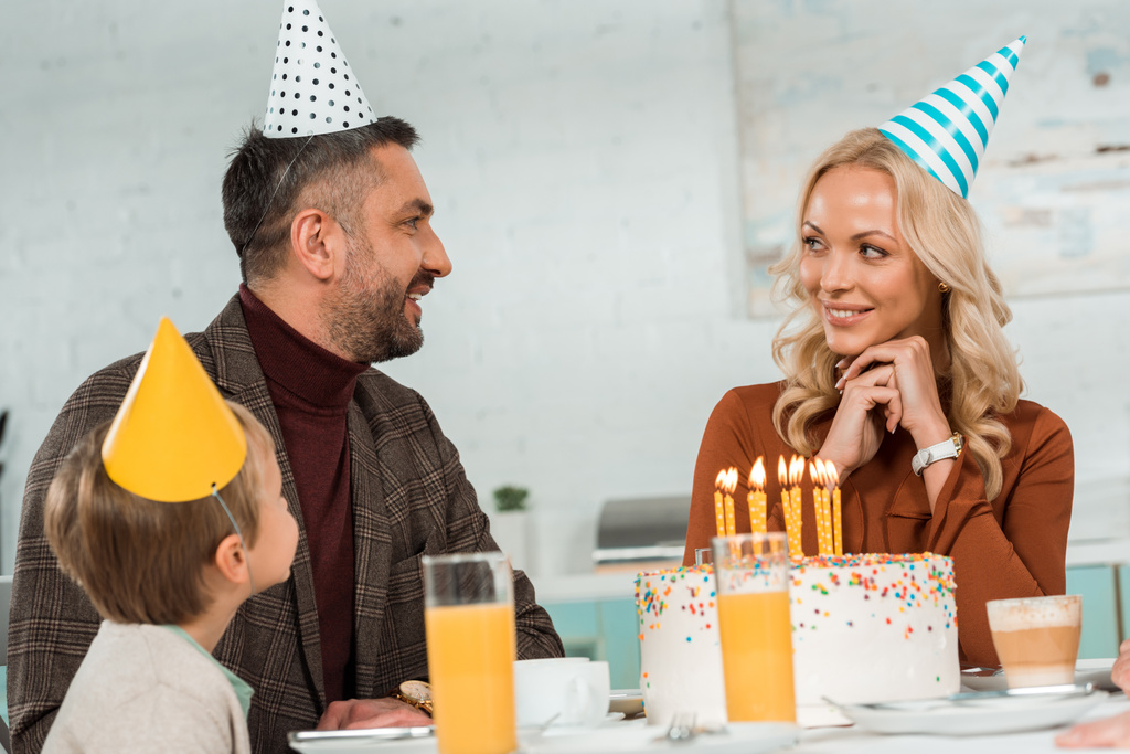 marido y esposa felices mirándose mientras están sentados en la mesa de la cocina con su hijo cerca del pastel de cumpleaños
 - Foto, imagen