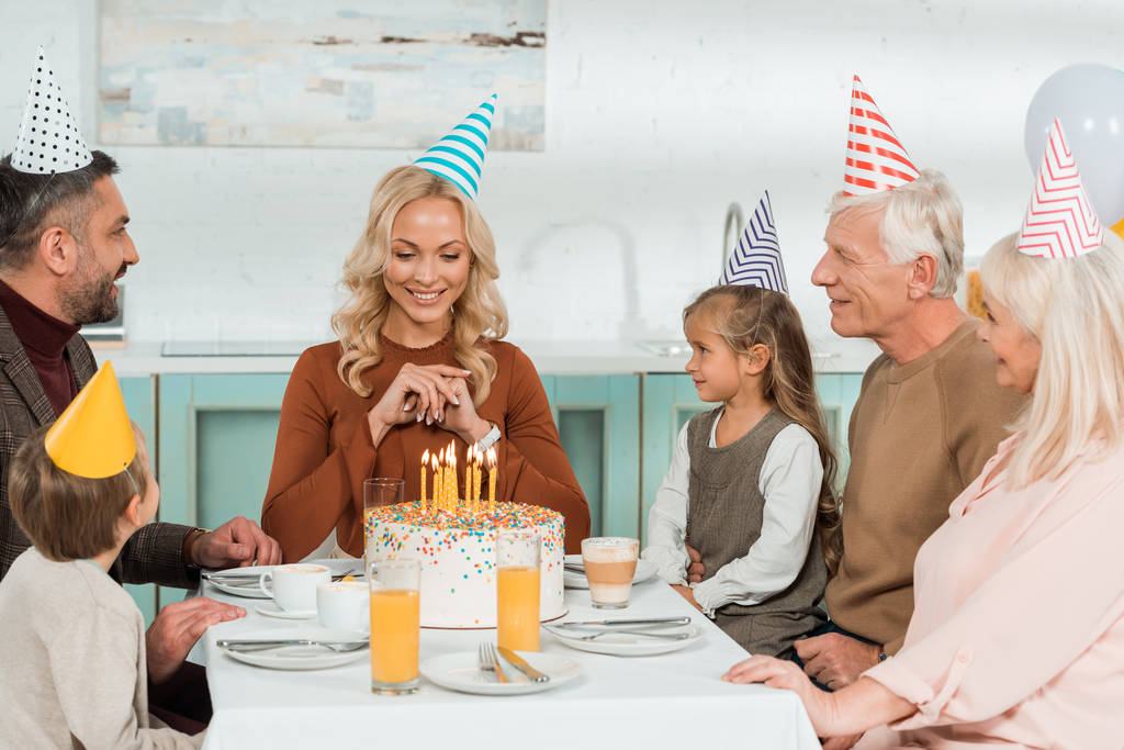 šťastná žena při pohledu na narozeninový dort, zatímco sedí u kuchyňského stolu s rodinou - Fotografie, Obrázek