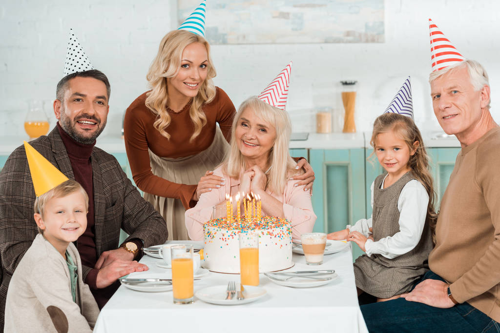 Улыбающаяся женщина касается плеч счастливой пожилой женщины, в то время как вся семья сидит возле торта день рождения
 - Фото, изображение