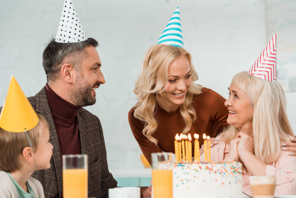 весёлая семья смотрит на счастливую пожилую женщину, сидящую рядом с праздничным тортом
 - Фото, изображение