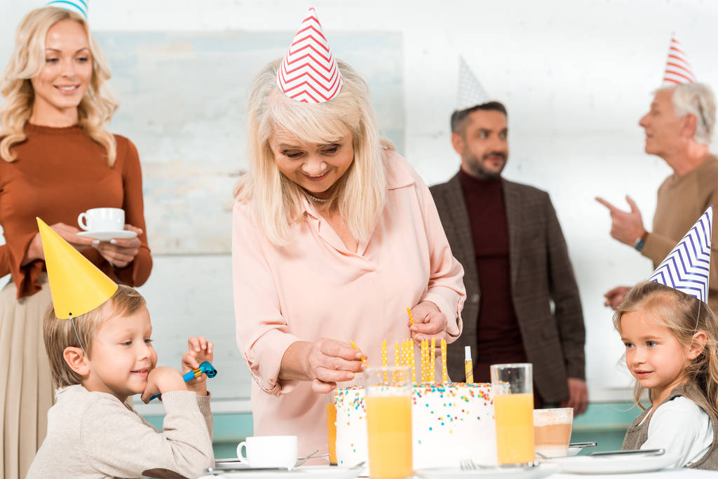 happy senior woman putting candles in birthday cake near family in party caps - Photo, Image