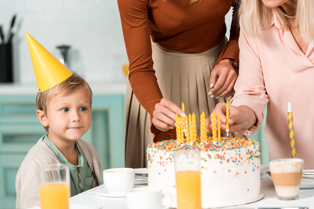 mignon garçon dans la casquette de fête en regardant mère et mamie mettre des bougies dans le gâteau d'anniversaire
 - Photo, image