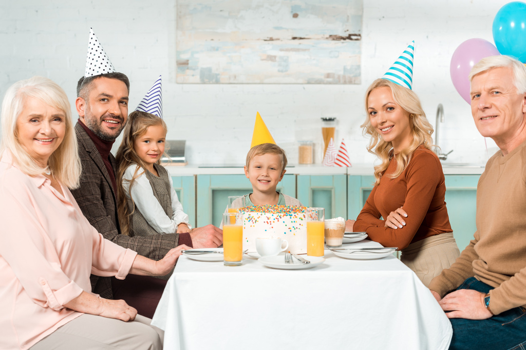 Mutlu bir aile mutfak masasında oturmuş doğum günü pastası yiyor ve kameraya bakıyor. - Fotoğraf, Görsel