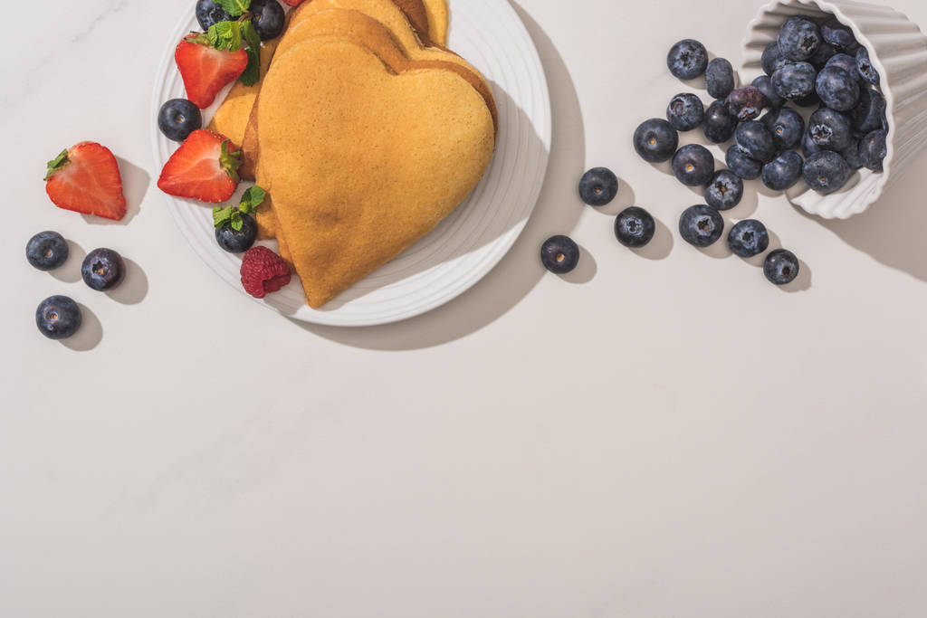 Draufsicht auf köstliche herzförmige Pfannkuchen in der Nähe von verstreuten Blaubeeren auf weißem Hintergrund - Foto, Bild