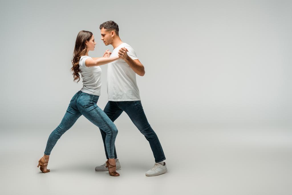 Seitenansicht von Tänzern in T-Shirts und Jeans, die Bachata auf grauem Hintergrund tanzen  - Foto, Bild