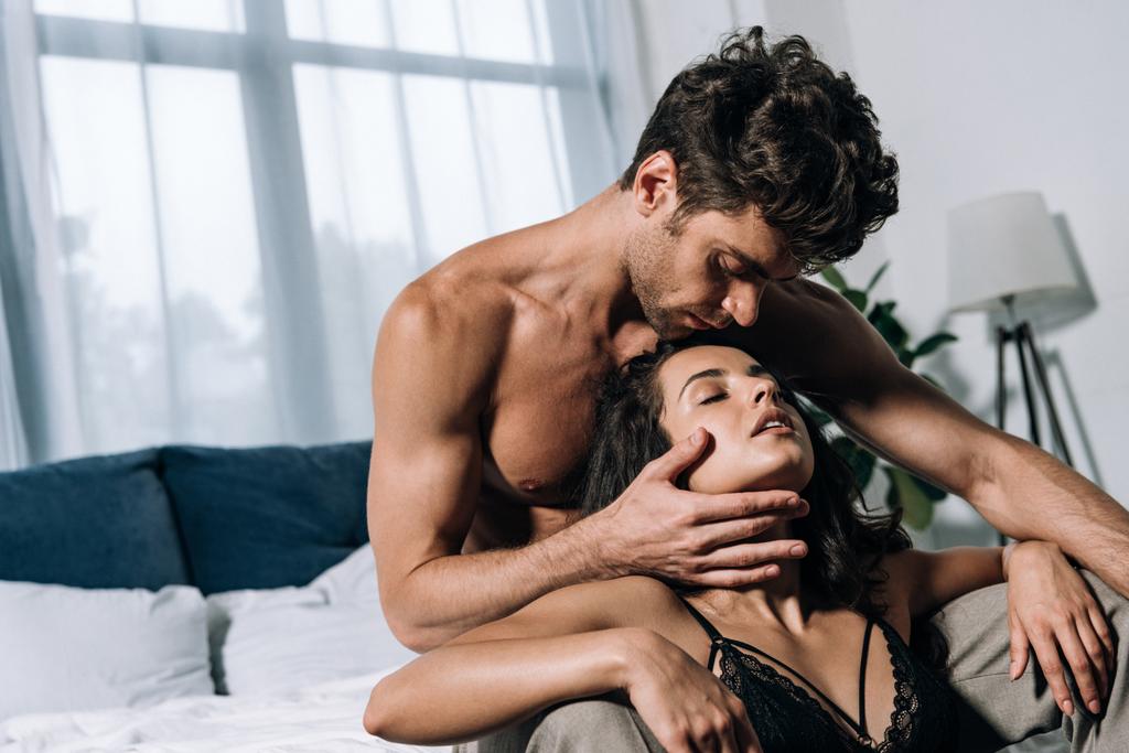 sensuele, shirtloze man zitten op bed en aanraken gezicht van sexy vriendin - Foto, afbeelding