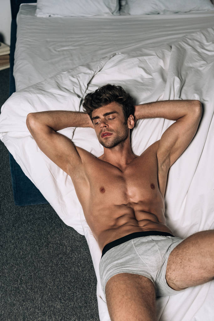 sexy, sem camisa homem olhando para longe enquanto deitado no chão perto da cama
 - Foto, Imagem