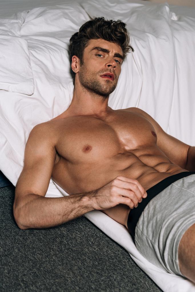sexy, hombre pensativo mirando a la cámara mientras está acostado en el suelo cerca de la cama
 - Foto, imagen
