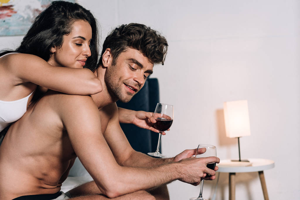 Mutlu genç çift yatak odasında kırmızı şarap bardakları tutuyor. - Fotoğraf, Görsel