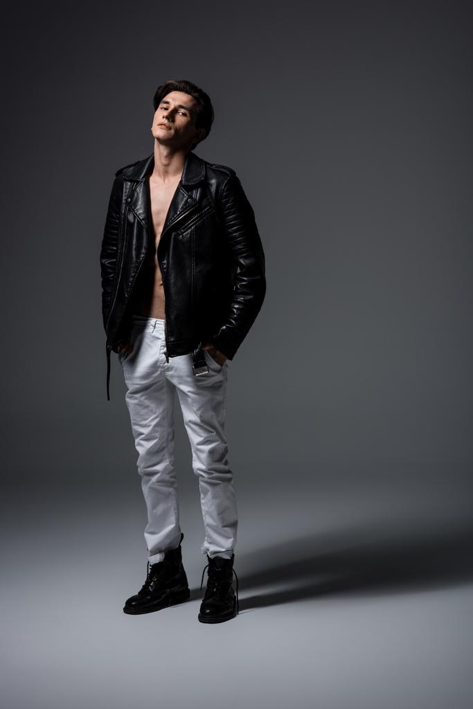 όμορφος σέξι άντρας με λευκό τζιν και μαύρο δερμάτινο μπουφάν, σε γκρι - Φωτογραφία, εικόνα