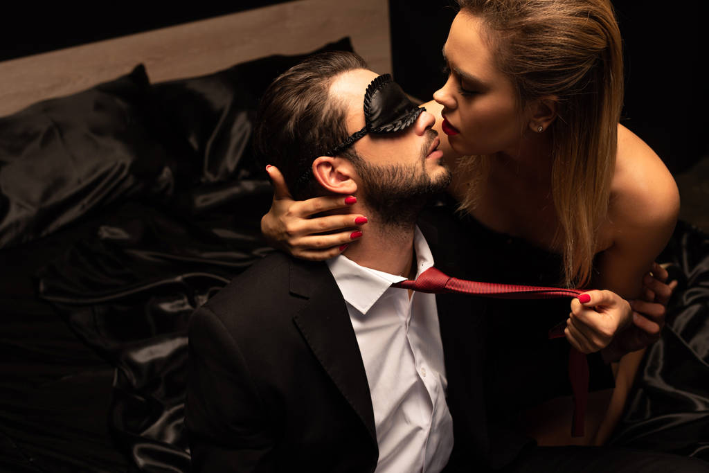 schöne sexy Frau küsst Mann in Maske auf Bett in dunklen Raum - Foto, Bild