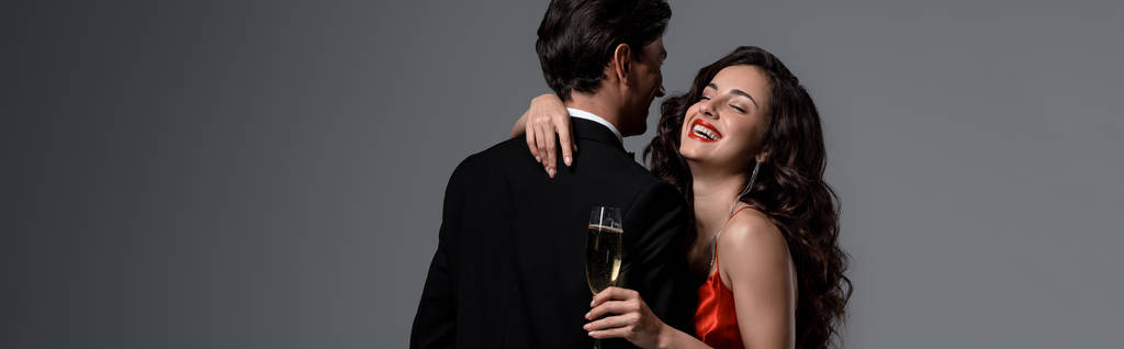 panoramic shot of beautiful smiling girl hugging boyfriend while holding glass of champagne, isolated on grey - Photo, Image