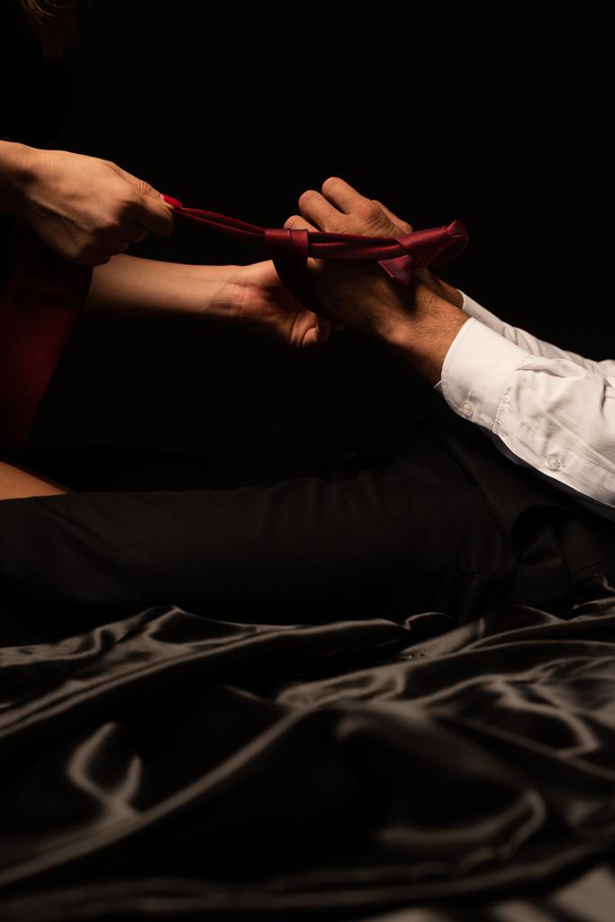 黒いベッドの上で男の手とタイを結ぶ女性のクロップドビュー - 写真・画像