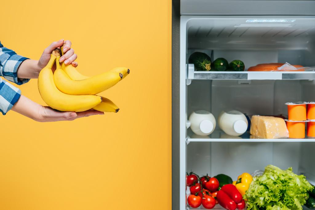 καλλιεργούμενη θέα της γυναίκας που κρατά μπανάνες κοντά σε ανοιχτό ψυγείο με φρέσκα τρόφιμα στα ράφια που απομονώνονται στο κίτρινο  - Φωτογραφία, εικόνα