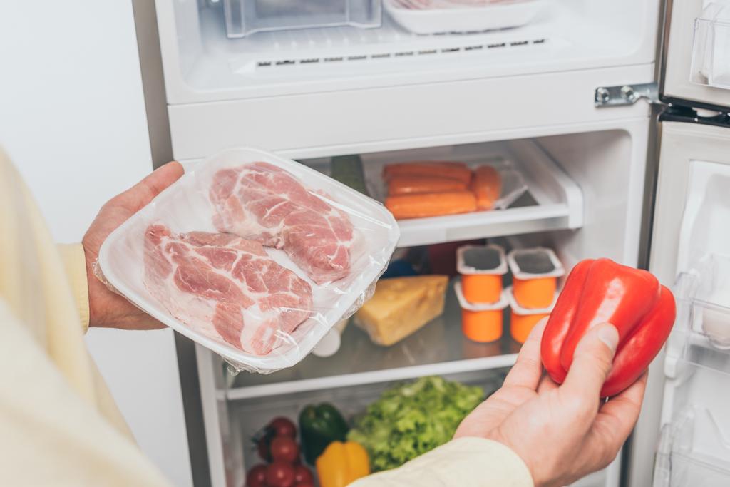 冷凍肉と新鮮なピーマンを食べ物でいっぱいのオープン冷蔵庫の近くに保持する男のクロップドビュー - 写真・画像