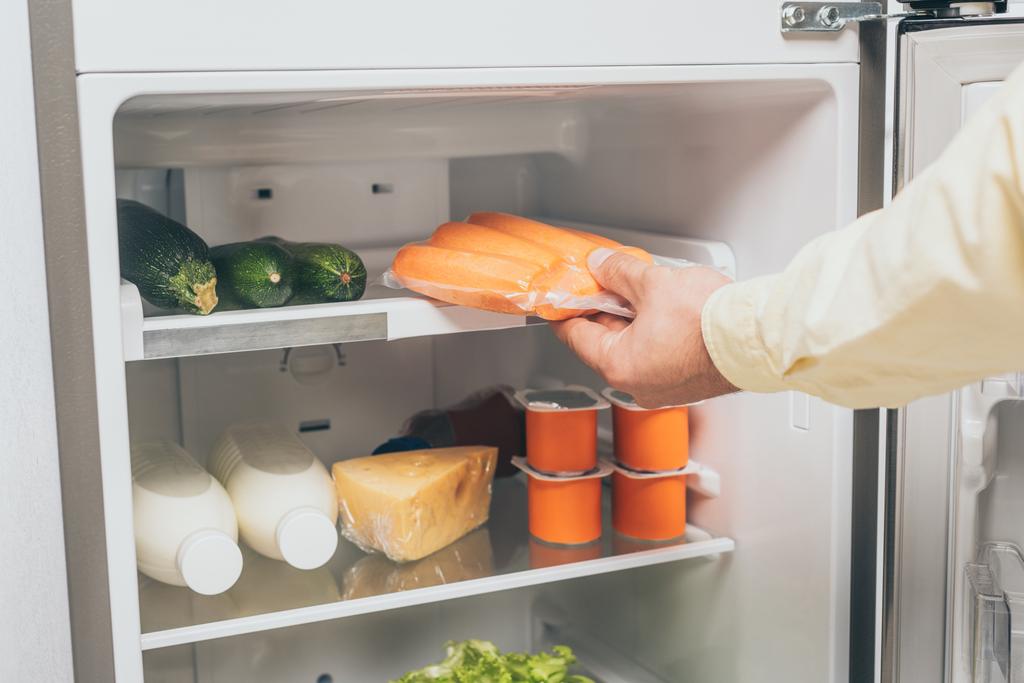 καλλιεργημένη άποψη του ανθρώπου που κρατά καρότα σε συσκευασία κενού κοντά σε ανοιχτό ψυγείο γεμάτο τρόφιμα - Φωτογραφία, εικόνα