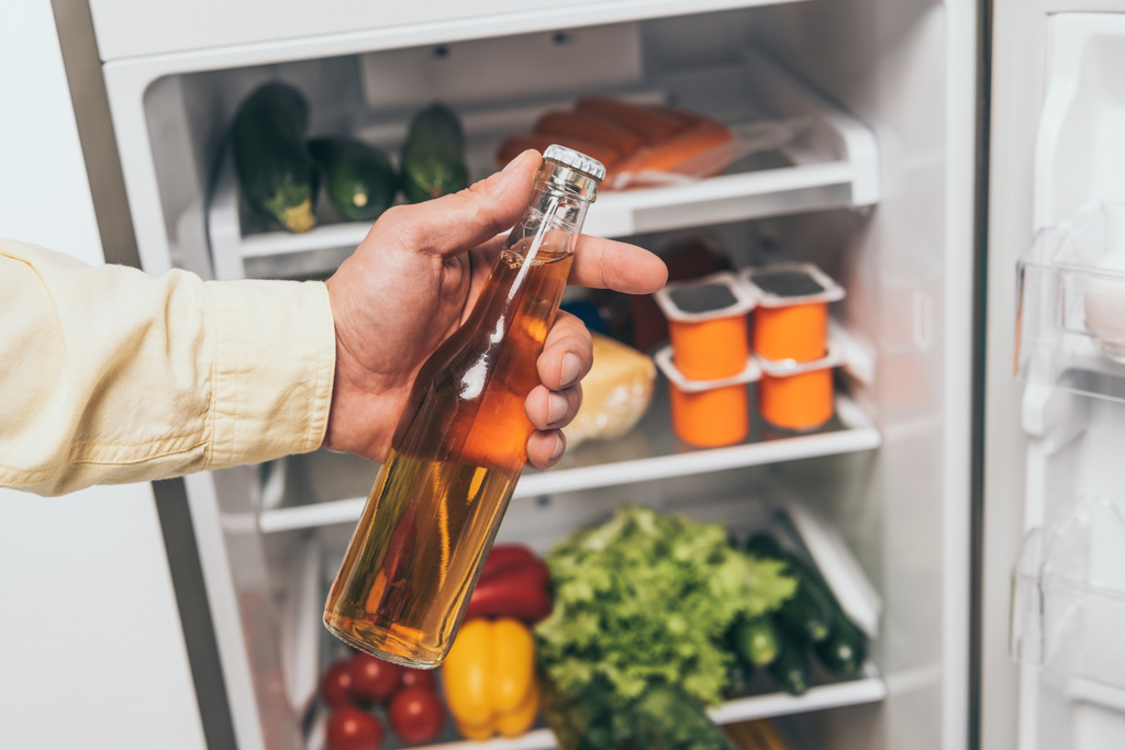 levágott kilátás férfi kezében üveg sör közelében nyitott hűtőszekrény tele étellel - Fotó, kép