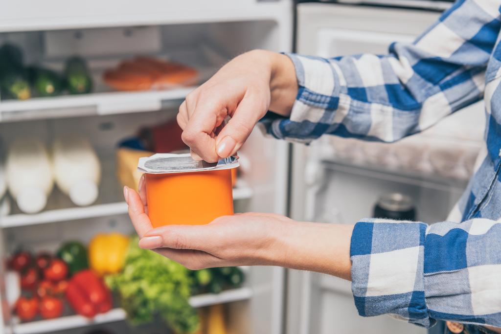 abgeschnittene Ansicht einer Frau, die Joghurt in der Nähe des offenen Kühlschranks mit frischen Lebensmitteln in den Regalen öffnet - Foto, Bild