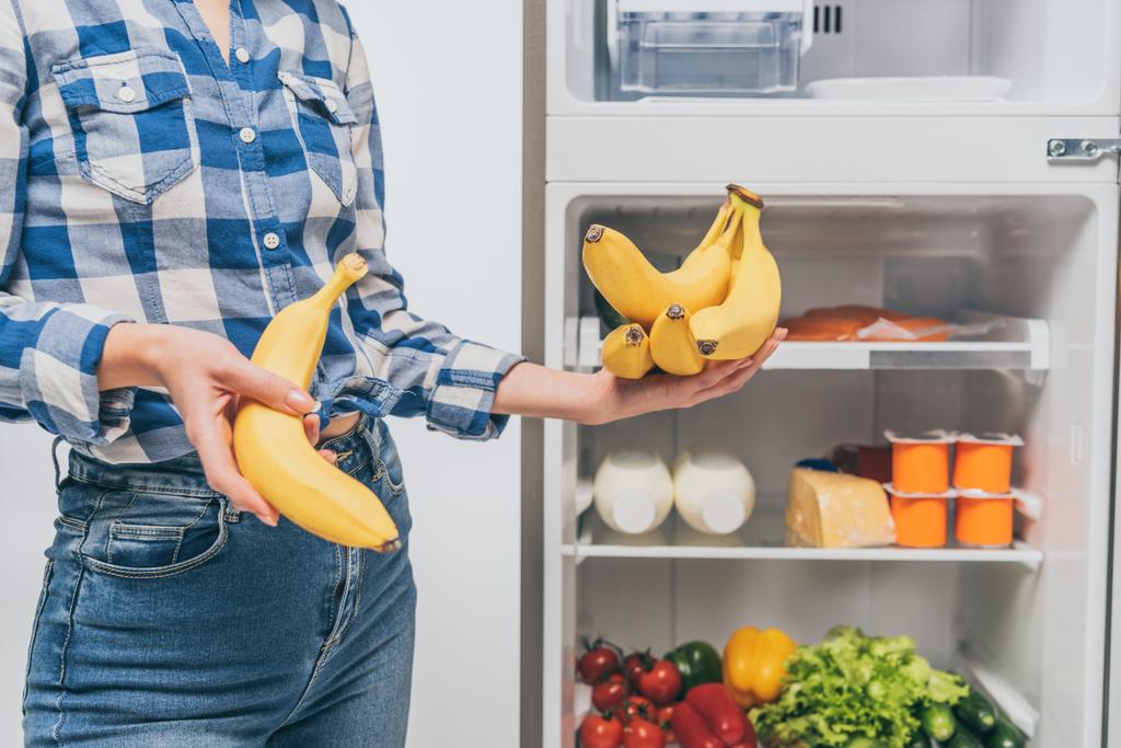 cropped view of woman holding bananas near open fridge with fresh food on shelves isolated on white - Photo, Image