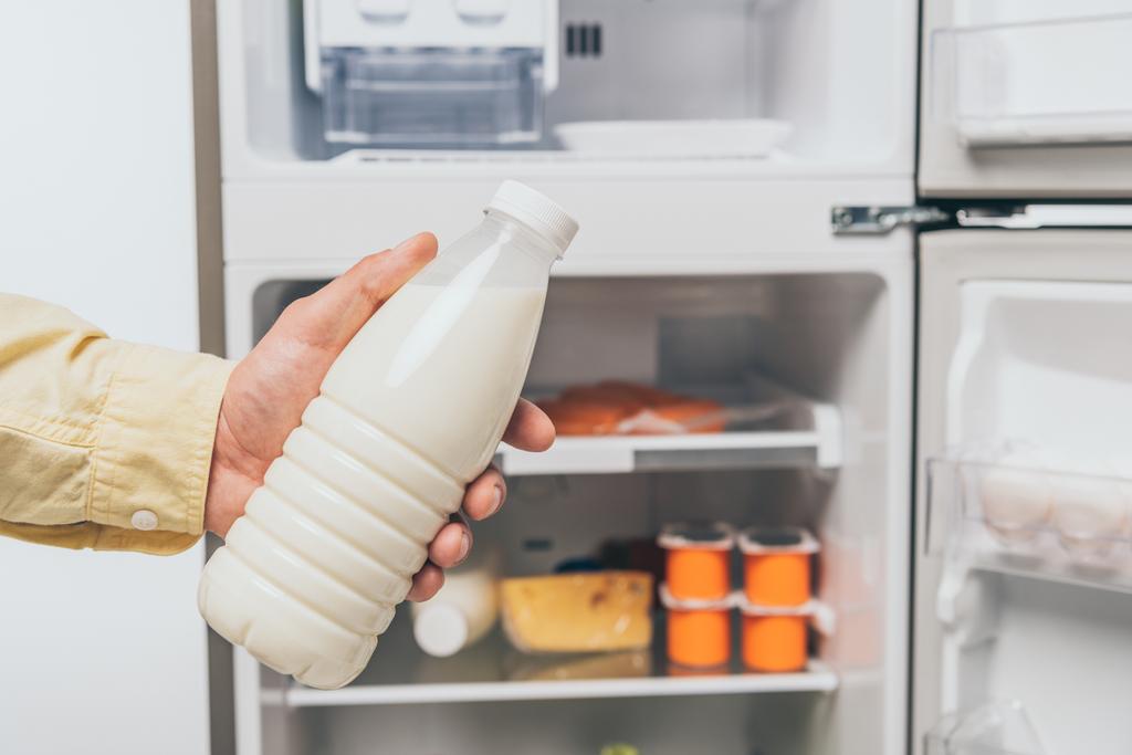 cropped view of man holding bottle of milk near open fridge with fresh food on shelves isolated on white - Photo, Image