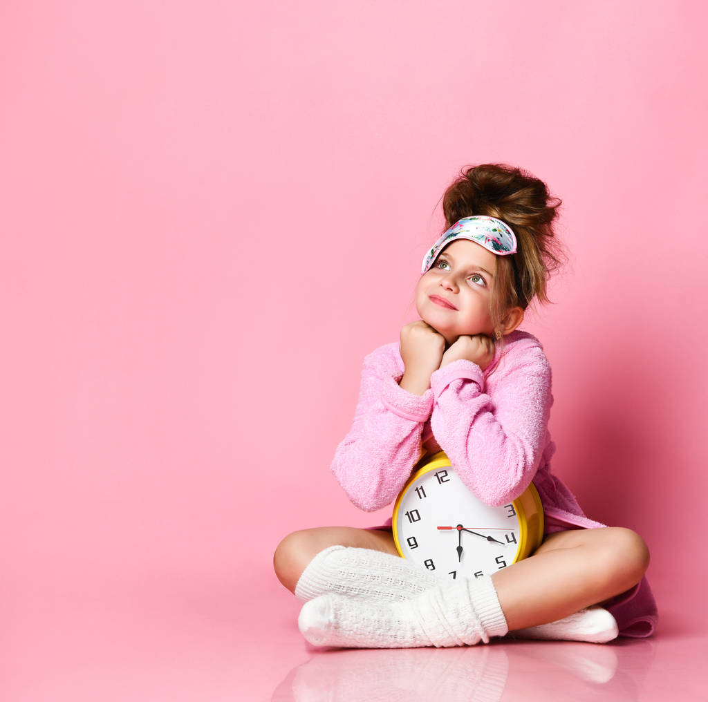 Χασμουρητό έφηβο κορίτσι κρατώντας ένα ξυπνητήρι, ενώ κάθεται σε μια housecoat στο πάτωμα. - Φωτογραφία, εικόνα