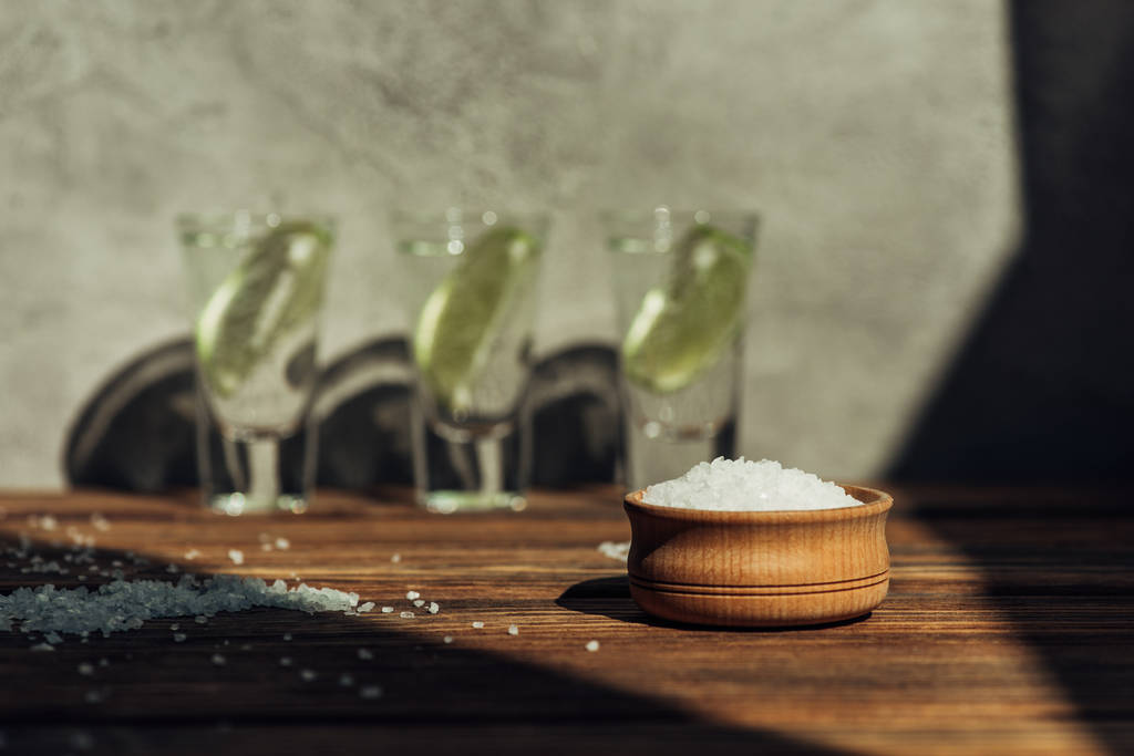 selektiver Salzfokus in Schüssel und frischer Tequila mit Kalk auf Holzoberfläche im Sonnenlicht - Foto, Bild