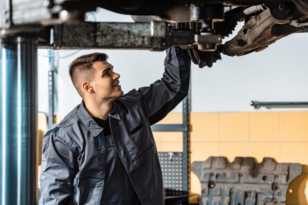 joven mecánico inspeccionando coche levantado en taller
 - Foto, Imagen