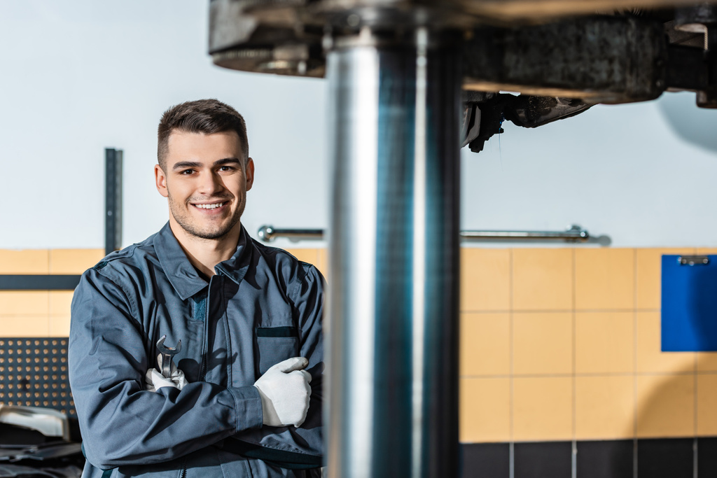 улыбающийся механик смотрит в камеру, стоя рядом с поднятой машиной в мастерской
 - Фото, изображение