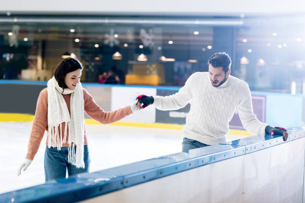 Χαρούμενη νεαρή γυναίκα διδάσκει τον άντρα να κάνει πατινάζ σε ένα παγοδρόμιο κρατώντας τα χέρια - Φωτογραφία, εικόνα