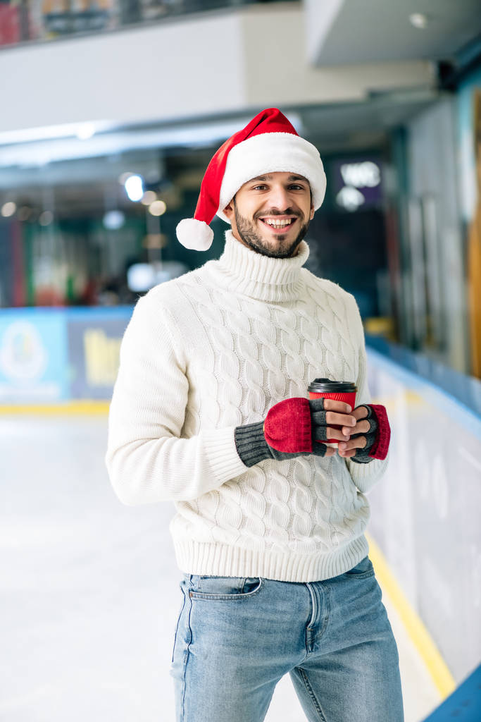 handsome cheerful man in sweater and santa hat holding coffee to go on skating rink - Photo, Image