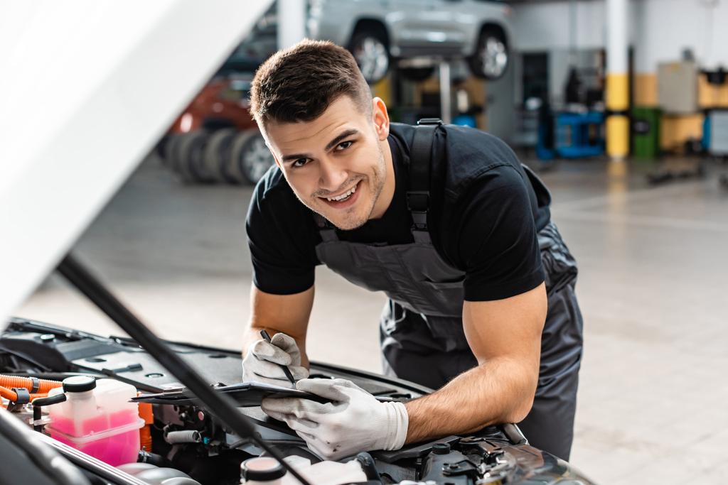 selektiver Fokus des lächelnden Mechanikers, der auf Klemmbrett schreibt, während er den Motorraum inspiziert - Foto, Bild