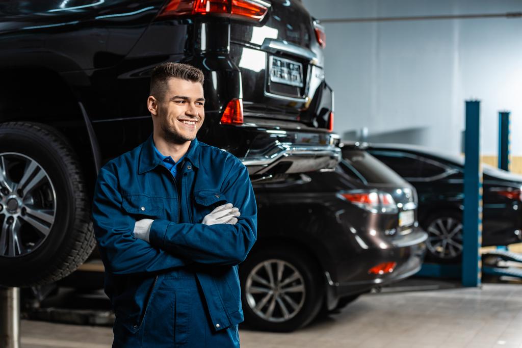 молодой, улыбающийся механик, стоящий со скрещенными руками и смотрящий в сторону современных автомобилей в мастерской
 - Фото, изображение