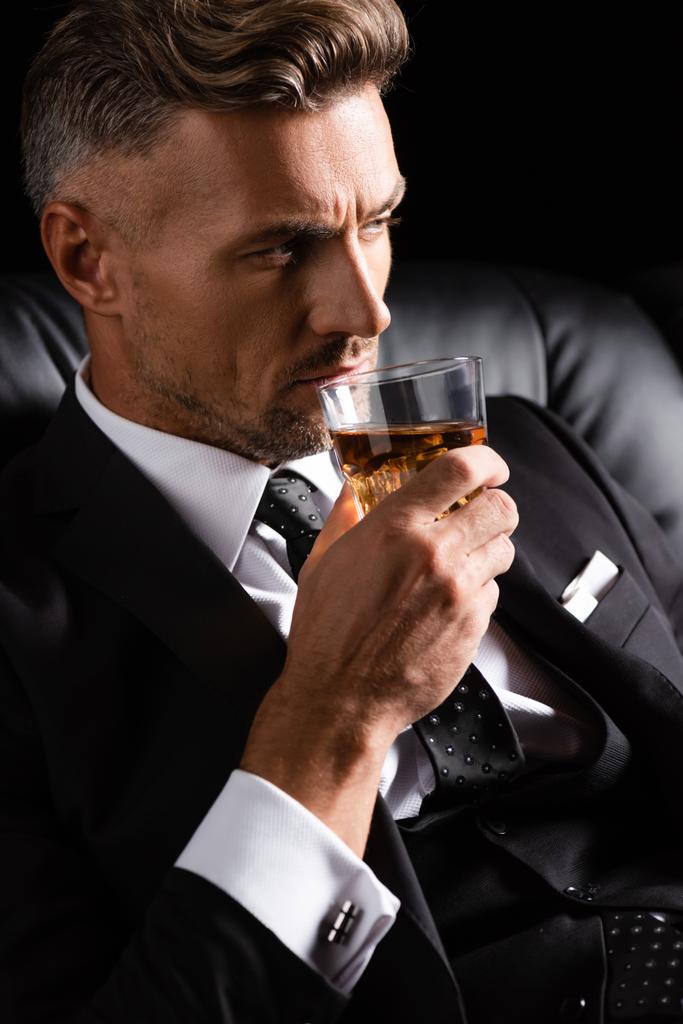 Empresário confiante em terno com copo de uísque no sofá isolado em preto
 - Foto, Imagem