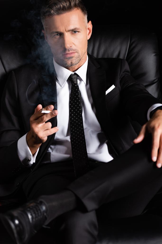 Selbstbewusster Geschäftsmann blickt in die Kamera und raucht Zigarette auf Sofa isoliert auf Schwarz  - Foto, Bild