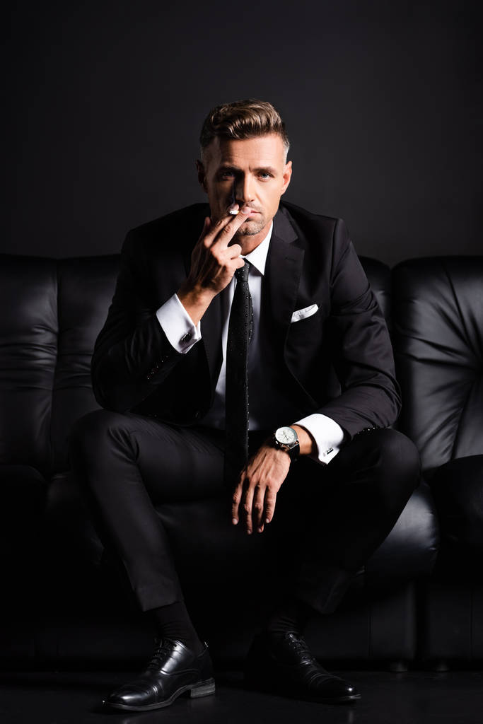 Empresario mirando a la cámara mientras fuma cigarrillo en sofá aislado en negro
 - Foto, Imagen