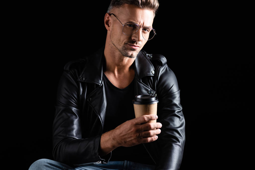 Stylish man in biker jacket looking at camera while holding disposable cup isolated on black  - Photo, Image
