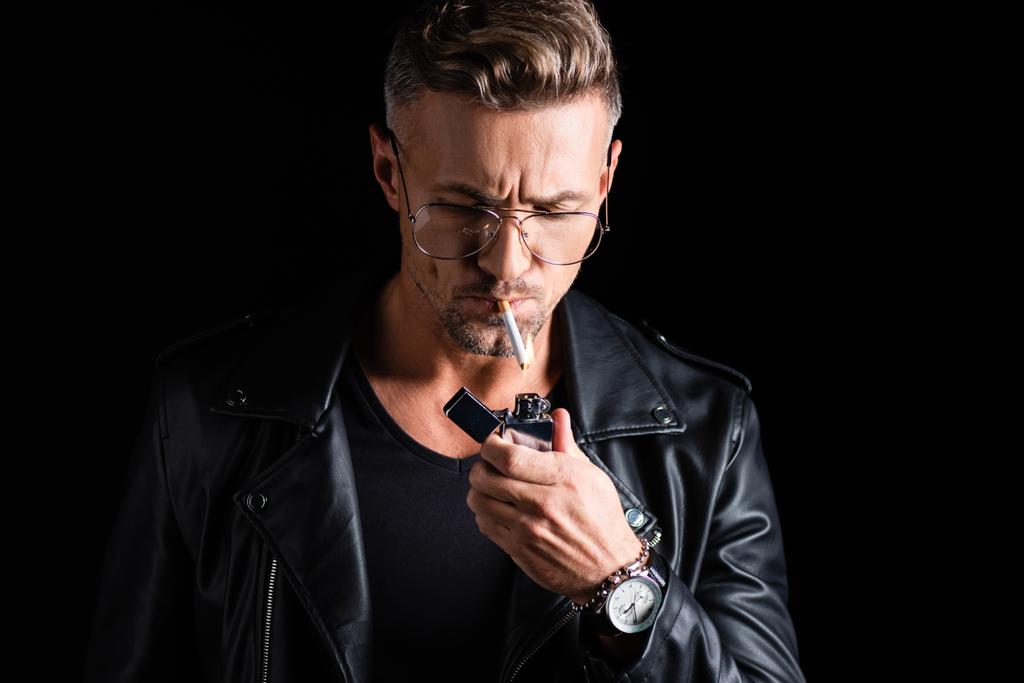 Handsome man in leather jacket lighting cigarette with lighter isolated on black - Photo, Image