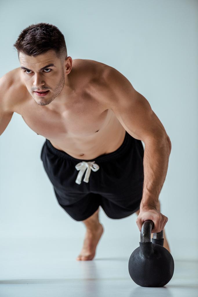 sexy gespierde bodybuilder met kale romp doen push-ups met kettlebell op grijs - Foto, afbeelding