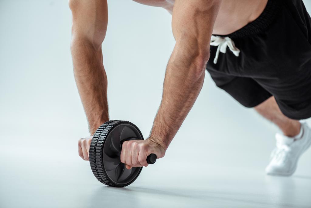 Ausgeschnittene Ansicht eines sexy muskulösen Bodybuilders mit nacktem Oberkörper, der mit Bauchtrainer auf grauem Hintergrund trainiert - Foto, Bild