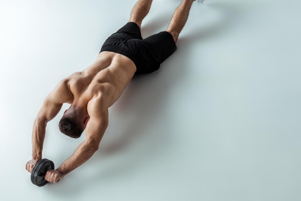 κάτοψη σέξι μυϊκή bodybuilder με γυμνό κορμό άσκηση με τροχό ab σε γκρι φόντο - Φωτογραφία, εικόνα