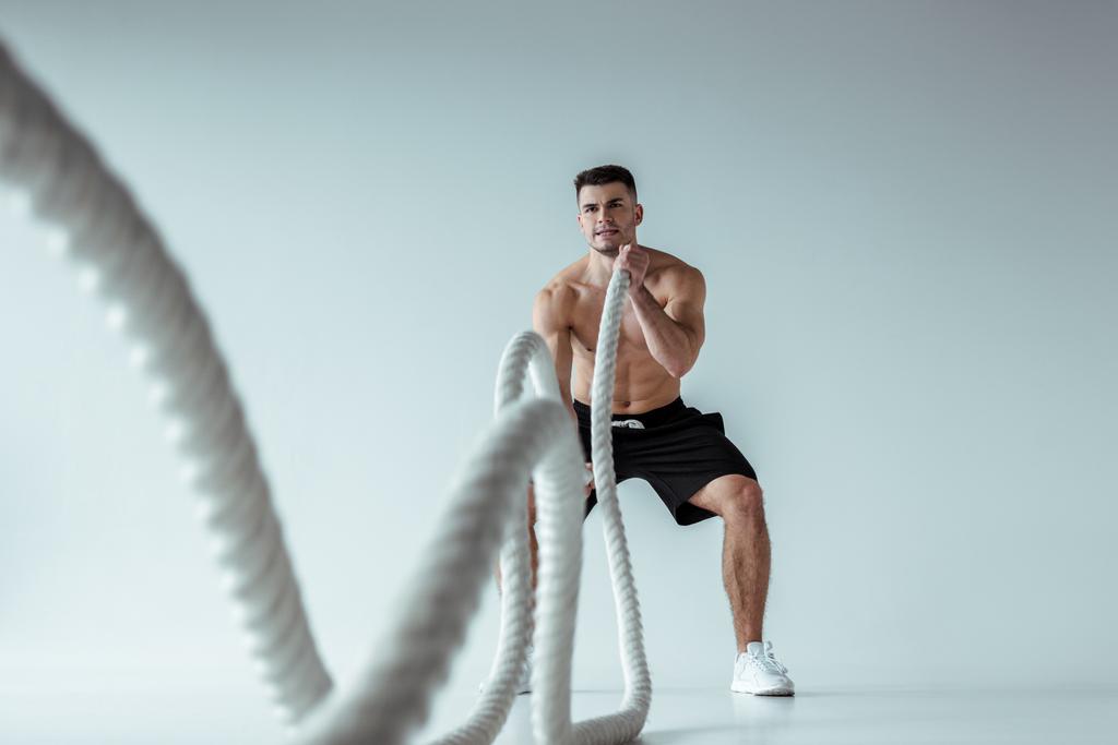 Selektiver Fokus der sexy muskulösen Bodybuilderin mit nacktem Oberkörper beim Training mit dem Kampfseil auf grauem Hintergrund - Foto, Bild