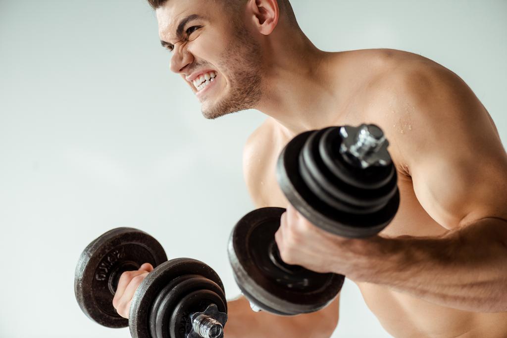 angespannter muskulöser Bodybuilder mit nacktem Oberkörper, der mit auf grau isolierten Kurzhanteln trainiert - Foto, Bild
