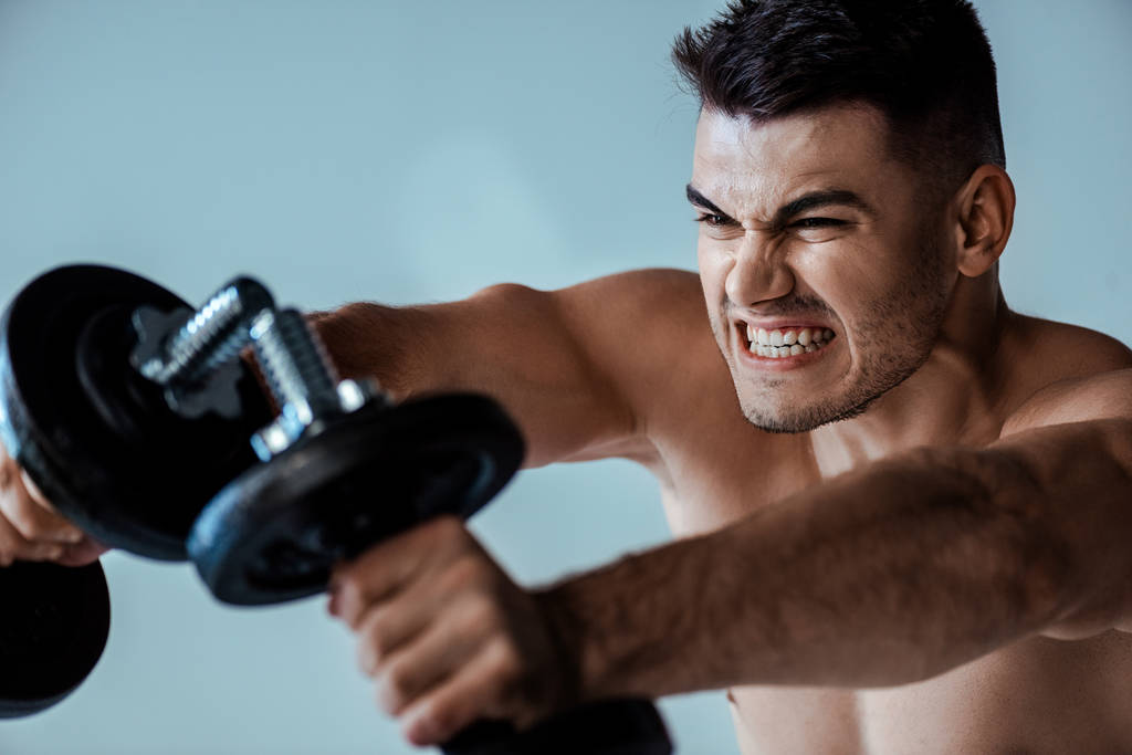 selective focus of tense muscular bodybuilder with bare torso working out with dumbbells isolated on grey - Photo, Image