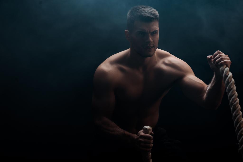 fisiculturista muscular sexy com tronco nu extirpando com corda de batalha no fundo preto com fumaça
  - Foto, Imagem