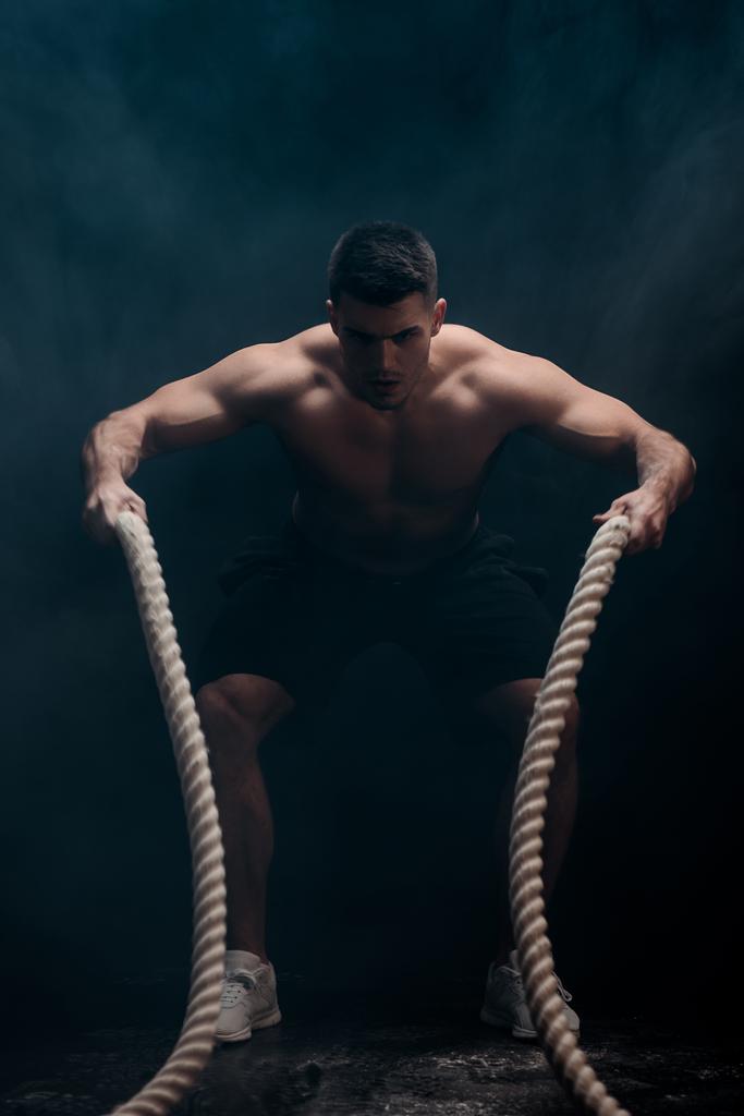 fisiculturista muscular sexy com tronco nu extirpando com corda de batalha no fundo preto com fumaça
  - Foto, Imagem